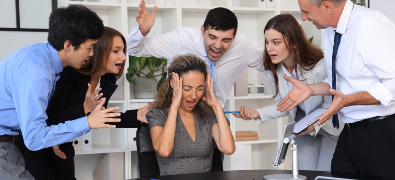 Acoso laboral. Mujer rodeada de otras personas que le increpan y tiene las manos sobre los oídos y cara de angustia