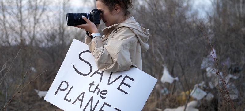 Joven con una cámara en la mano haciendo una imagen de una zona con basura y un cartel en un lateral en el que se lee save the planet (salvar el planeta)