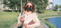 Chica que pasea por un parque con libros, café, gafas de sol  y mascarilla.
